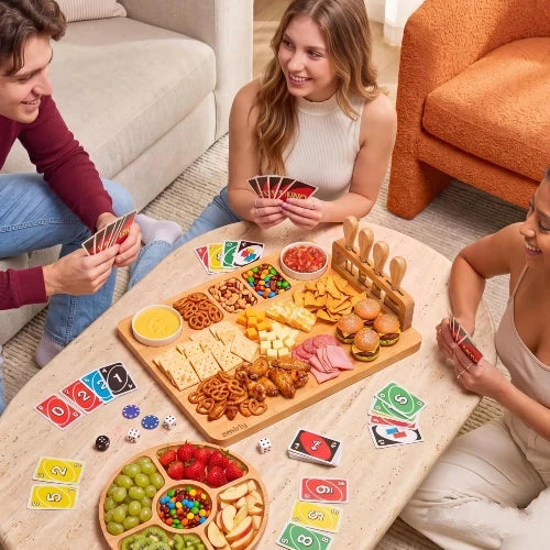 Friends playing a card board game around a living room coffee table enjoying snacks and sliders on a NovoBam bamboo charcuterie platter with round fruit tray