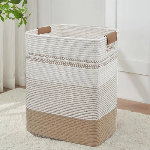 Tall rectangular woven cotton rope laundry hamper in jute and white color-block design with leather handles, standing on a soft rug in a clean room.