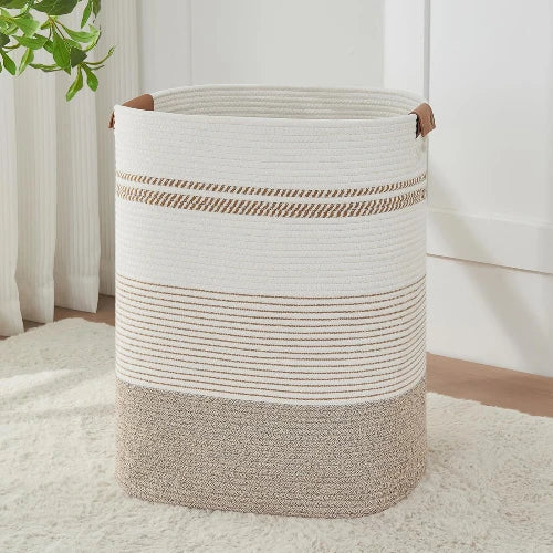 Tall rectangular woven cotton rope laundry hamper in light brown and white stripes with leather handles, standing upright in a bedroom.