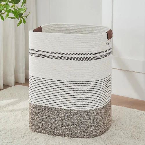 Tall rectangular woven cotton rope laundry hamper in rice brown and white stripes with leather handles, standing upright in a bedroom.
